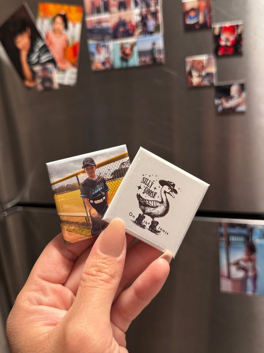 Hand holding a small square magnet with a design on a refrigerator covered with other magnets.